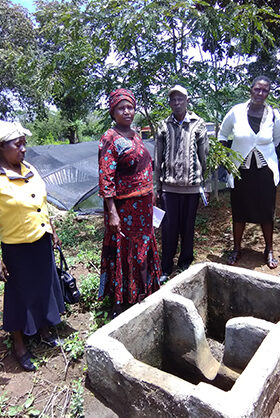 Mary (in red) with leaders of Makakya Community Based Organisation. In front of them is the drainage tub which feeds water into the pond. Behind them, covered with a large plastic net, is the pond.